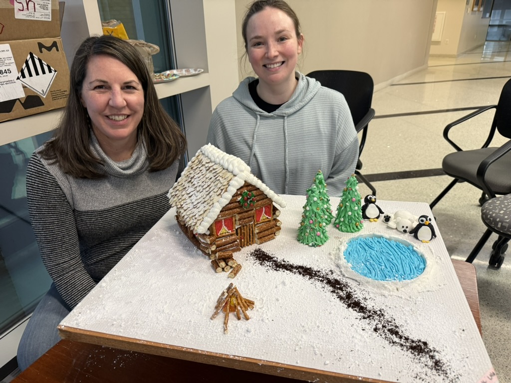 Professor and graduate student posing next to gingerbread house they decorated. 