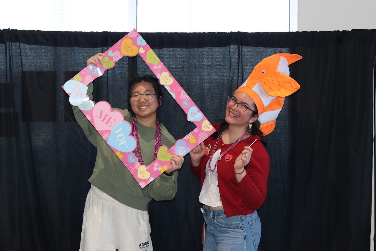 Two students posing with a heart frame and fish hat. 